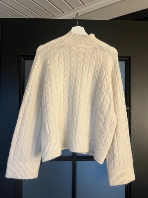 Vit stickad tröja från Carin Wester - Säljer en vit stickad tröja från Carin Wester i storlek M. Tröjan har ett klassiskt flätmönster, hög ribbad hals och breda muddar vid ärmslut och nederkant. Perfekt för kyliga dagar och riktigt mysig att bära.