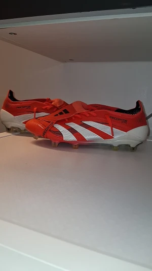 Adidas Predator Elite fotbollsskor röd/vit - Säljer ett par Adidas Predator Elite fotbollsskor i rött och vitt med svarta detaljer. Skorna har snörning, mönstrad sula med dobbar för gräs och klassisk Predator-logga undertill. Ovandelen är i syntetmaterial och innersulan är limegrön med svart grafik. Perfekta för dig som vill sticka ut på planen. 