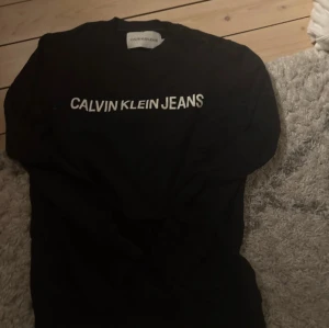 Svart långärmad t-shirt Calvin Klein Jeans - Svart långärmad t-shirt från Calvin Klein Jeans i storlek XS. Klassisk rund hals och vit logotyp tryckt framtill. Mjuk och skön bomullskvalitet som passar perfekt till jeans eller shorts. Enkel och stilren design med normal passform. Knappt använd 
