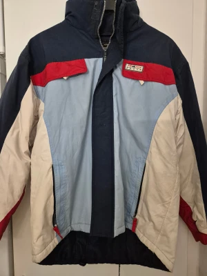 Skidjacka/Vinterjacka - Snygg retro skidjacka/vinterjacka från Gianfranco i storlek S med färgblock i marinblått, ljusblått, rött och vitt. Jackan har hög krage, huva och dragkedja framtill. Perfekt för kalla dagar och har en sportig vibe med FR Snow Sport-detalj på bröstet. Har några fläckar.