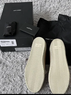 Saint Laurent beige sneakers mocka - Snygga beige sneakers från Saint Laurent med handskriven logga på sidan. Skorna har vit platt sula, rund tå och vita snören. Tillverkade i mjuk mocka med vita detaljer vid hälen. Perfekta för dig som gillar stilrena och exklusiva sneakers.