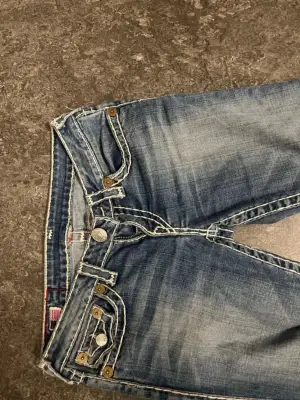 Säljer ett par blå jeans från True Religion med tydliga vita kontrastsömmar och ikoniska fickdetaljer. Låg midja och raka ben, klassisk femficksmodell med silverfärgade knappar och logopatch bak. Perfekta för dig som gillar street och statement.