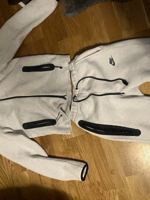 Nike tech fleece  - Nike mjukisbyxor i ljusgrått med svarta dragkedjor och snörning i midjan. Byxorna har en clean look med Nike-logga på låret och praktiska fickor med svarta zippar. Perfekt för chill eller träning, tillverkade i mjukt bomullsmaterial.
