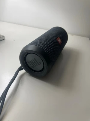 JBL Flip 4 - JBL Flip 4 bärbar Bluetooth-högtalare i svart. Ger kraftfullt ljud och har en robust, vattentålig design. Högtalaren ser ut att vara i gott skick utan synliga skador eller slitage. Perfekt för musik både hemma och utomhus.