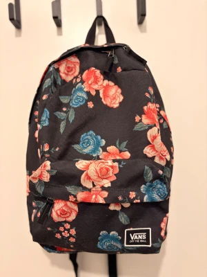 Blommig ryggsäck från Vans - Svart ryggsäck från Vans med stort blommönster i rosa och blå toner. Ryggsäcken har en klassisk form med justerbara axelremmar, dragkedja och en praktisk ytterficka framtill. Perfekt för skolan eller stan och ger en färgstark touch till din outfit.