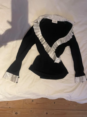 Svart tröja med prickiga detaljer  - Svart långärmad tröja från Wallis med vit krage, manschetter och volangkant i satin med svarta prickar. Snygg v-ringning och dubbla knappar på ärmsluten. Figurnära passform. På storlekslappen står det 12 petite, vilket motsvarar typ strl M.