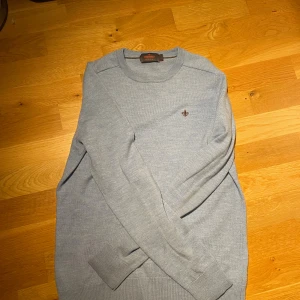 Ljusblå merinoulltröja från Morris - Snygg ljusblå tröja från Morris i 100% merinoull. Klassisk rund hals och diskret Morris-logga broderad på bröstet. Tröjan har långa ärmar och ribbade muddar vid ärmslut och nederkant. Perfekt för dig som gillar stilrena och bekväma plagg. Storlek M men passar S