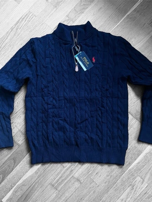 Marinblå stickad tröja Polo Ralph Lauren - Snygg marinblå stickad tröja från Polo Ralph Lauren med kabelstickat mönster och röd logobroderi på bröstet. Tröjan har half zip-dragkedja, ribbad krage och långa ärmar. Perfekt för dig som gillar klassisk stil med en modern touch.