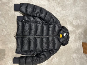 Svart pufferjacka med huva - Svart pufferjacka från Parajumpers med huva och ribbade muddar. Jackan har quiltad design, dragkedja framtill och praktiska fickor med dragkedja. Materialet är glansig polyester och jackan har en gul detalj vid halsen. Perfekt för kalla dagar.