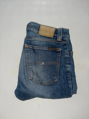 Nudie Jeans  - Säljer ett par Nudie Jeans Tube Kelly i klassisk blå tvätt. Jeansen har raka ben och snygga kontrastsömmar. Tillverkade i ekologisk bomull med lite stretch för extra komfort. Midja 37cm, längd 108cm. JAG SKICKAR EJ FLER BILDER NÄR JAG BÄR JEANSEN! för storleksguide hänvisar jag till måtten och för bättre inblick i passformen rekommenderar jag att googla på modellnamnet. kolla måtten noggrant :) s5 
