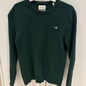 Mörkgrön ulltröja från GANT - Säljer en mörkgrön stickad tröja från GANT i extra fin lammull. Tröjan har rund hals, lång ärm och en liten broderad logga på bröstet. 