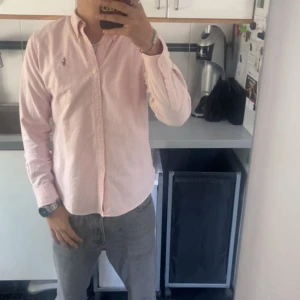 Rosa randig skjorta från Ralph Lauren - Snygg rosa och vit randig skjorta från Ralph Lauren med klassisk krage och broderad logga på bröstet. Skjortan har långa ärmar, knappar framtill och är gjord i mjuk bomull. Perfekt för dig som gillar stilrena och fräscha plagg.