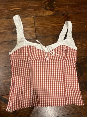 Rutig korsett-topp zara - Säljer en söt korsett-topp med breda, rynkade axelband och snörning framtill. Toppen har ett rödvitt rutigt mönster och vit överdel. Materialet känns lätt och somrigt, perfekt för att styla med jeans eller kjol. Passformen är normal och toppen är ärmlös. storlek S