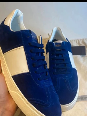 Valentino Garavani blå sneakers strl 44 - Säljer ett par snygga Valentino Garavani sneakers i blå mocka med vita skinnpaneler och vit platt sula. Skorna har snörning och coolt mönstrad yttersula. Perfekt för dig som gillar exklusiva och stilrena sneakers med unik design. MÖTS BARA ‼️