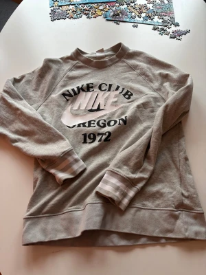 Grå sweatshirt från Nike Club - Säljer en grå sweatshirt från Nike Club med tryck 'Oregon 1972' på bröstet. Tröjan har långa ärmar med ljusgrå och vita ränder vid mudden. Perfekt för chill dagar och sportiga outfits.