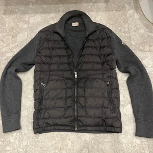 Moncler mörkgrå cardigan - har några hål på vissa ställen men inga som syns när man har på sig jackan. fråga om bilder. har kvitto Snygg mörkgrå pufferjacka från Moncler med quiltad framsida och stickade ärmar. Jackan har två dragkedjefickor, hög krage och Moncler-logga på ärmen. Materialmix av polyester och stickad ull ger både värme och stil. Perfekt för dig som vill ha en clean look.