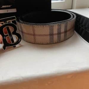 Beige rutig skärp från Burberry - Säljer ett snyggt Burberry-skärp med klassiskt rutig design i beige, svart och röd. Skärpet har en bred form och är tillverkat i skinn med en cool metallspänne med Burberrys logotyp. Perfekt för att lyfta din outfit med lite lyxig vibe.