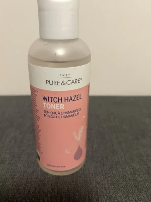 Witch Hazel Toner från Pure & Care - Fräsch ansiktstoner med trollhassel, ros och aloe vera som hjälper till att minimera porer, reglera talgproduktionen och bevara hudens fukt. Kommer i en genomskinlig plastflaska med rosa etikett och vitt lock. Perfekt för dig som vill ha glow och jämnare hudton.