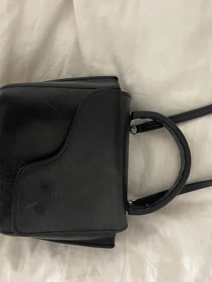 Svart axelväska från ATP ATELIER - Säljer en stilren svart axelväska i skinn med ett unikt lock och silverfärgade metalldetaljer. Väskan har både handtag och en lång axelrem som kan tas av med karbinhakar. Hyfsat bra skick men finns en hel del repor och liknade som man kan se på bilden 