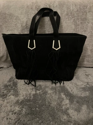 Bershka Svart väska med strängar  - Stor svart väska från Bershka i mocka med två rejäla handtag och snygga silverfärgade detaljer där handtagen möter väskan. Långa fransar hänger ner från handtagen och ger en cool touch. Perfekt för dig som gillar stilrena och trendiga accessoarer.