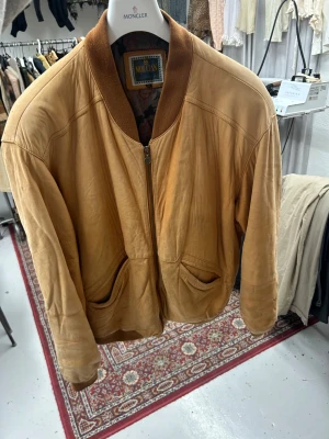 Beige skinn bomberjacka från Moutan - Snygg beige bomberjacka i skinn från Moutan med ribbad krage och muddar. Jackan har två framfickor och dragkedja framtill. Perfekt för dig som vill ha en klassisk och chill look med lite vintage-vibe och snygga slitningar och lös passform.
