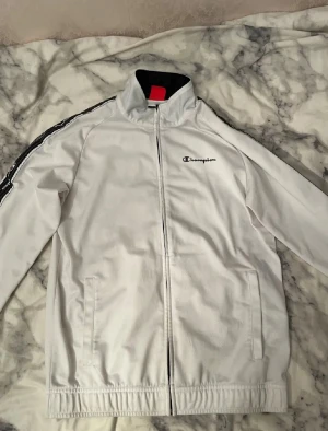 Vit champion tracksuit set - Bilderna visar ett vitt Champion track jacket och svarta Champion track pants, båda i polyester och storlek medium. Inga skor syns på bilderna.