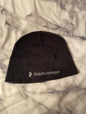Svart mössa från Peak Performance - Säljer en svart mössa från Peak Performance med broderad logga framtill. Mössan är ribbstickad och har en enkel, klassisk form som passar perfekt för kyliga dagar. Perfekt accessoar för dig som gillar stilren design.