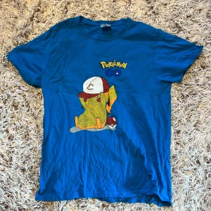 Blå Pokémon Go Pikachu t-shirt från Keya - Blå t-shirt från Keya med tryck av Pikachu i keps och Pokéball samt Pokémon Go-logga på bröstet. Klassisk rund hals och korta ärmar. Gjord i mjuk bomull, perfekt för dig som älskar Pokémon och vill sticka ut med en färgglad look. Trycket är inte i sitt bästa skick som man kan se (se bild 2) Storlek S