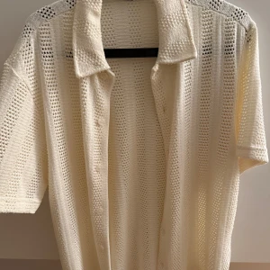 Vit stickad skjorta  - Snygg vit stickad skjorta med korta ärmar och klassisk krage. Skjortan har ett luftigt hålmönster i vertikala ränder och stängs med diskreta knappar framtill. Perfekt för dig som gillar en chill och unik stil. Använd kanske en gång, så i princip oanvänd! Mvh 