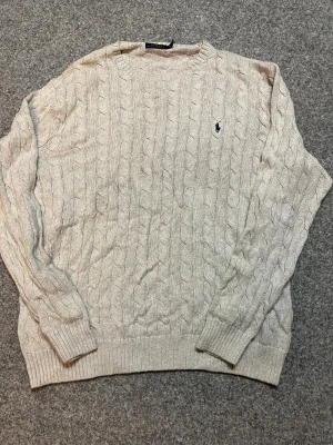 Beige kabelstickad tröja Polo Ralph Lauren - Säljer äkta beige kabelstickad tröja från Polo Ralph Lauren med klassisk blå logga broderad på bröstet. Tröjan har rund hals, ribbade muddar och är långärmad. Perfekt för dig som gillar stilrena och tidlösa plagg.