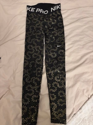 Nike Pro svarta leggings med guldmönster - Nike Pro leggings i svart med guldigt abstrakt mönster och prickar. Bred midja med vit Nike Pro-logga. Sitter tight och är perfekta för träning eller chill. Materialet är stretchigt syntet som andas och ger bra rörelsefrihet.