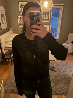 Svart Moncler cardigan - Säljer en svart pufferjacka från Moncler med stickade ärmar och quiltad framsida. Jackan har två fickor med knappstängning, rund hals och dragkedja framtill. Moncler-logga på ärmen ger en snygg detalj. Perfekt för dig som gillar stilrena och sportiga plagg.