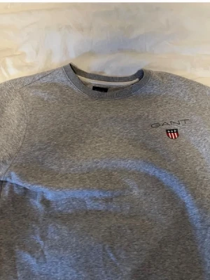Grå sweatshirt från GANT - Snygg grå sweatshirt från GANT med broderad logga och emblem på bröstet. Tröjan har rund hals, ribbade muddar och är långärmad. Perfekt för chill dagar och enkel att matcha med jeans eller joggers. Storlek M.