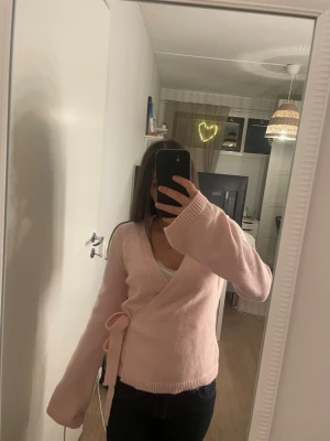 Ljusrosa omlottkofta från SHEIN - Säljer en ljusrosa stickad omlottkofta från SHEIN i storlek M. Koftan har långa ärmar, v-ringad hals och knytband i sidan för en snygg passform. Perfekt att slänga på sig när det är lite kyligt ute.