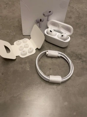 AirPods Pro 2 - Apple AirPods Pro 2 med laddningsetui, extra öronkuddar och laddningskabel. Produkten ser ut att vara i mycket gott skick utan synligt slitage. Aktiv brusreducering och trådlös anslutning för bästa ljudupplevelse. Perfekt för musik, samtal och träning.