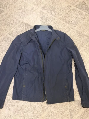 Blå vindjacka från Belstaff - Snygg marinblå vindjacka från Belstaff med klassisk ståkrage och dragkedja framtill. Jackan har långa ärmar, två fickor med tryckknappar och coolt Belstaff-märke på ärmen och insidan. Storlek är 16yr men passar S. Därav billiga priset. Jackan knappt använd och passar perfekt med hoodie under. PRIS kan diskuteras vid snabaffär