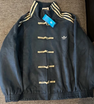 adidas limited chinese/lunar jacka - Adidas Originals vindjacka i mörkblått med beige snörning framtill och klassiska tre ränder på axlarna. Jackan har resår vid ärmslut och nederkant, samt två sidofickor. Materialet är polyester och jackan har lång ärm. Perfekt för dig som gillar sportig stil. JACKAN ÄR LIMITERADE VILKET ÄR SVÅRT ATT FÅ TAG PÅ
