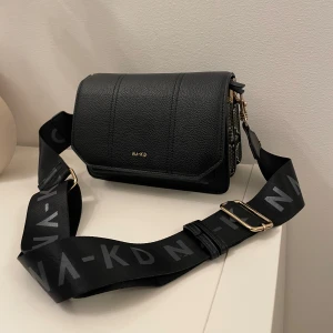 Svart axelväska från NA-KD - Säljer en svart axelväska från NA-KD med bred justerbar rem med logotyp. Väskan har en stilren design i skinnimitation och detaljer i guld. Sidorna har ett coolt ormmönster som ger extra edge. Perfekt storlek för det viktigaste och passar till många outfits.