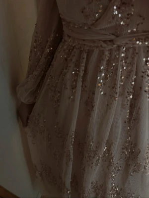 Lång ljus klänning med paljetter - Säljer en elegant långklänning i ljus beige ton med glittriga paljettdetaljer över hela tyget. Klänningen har långärmade ärmar, omlottliv och ett knytband i midjan. Materialet är lätt och skirt, perfekt för speciella tillfällen.