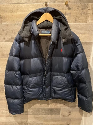 Polo Ralph Laurent jacka  - Snygg mörkblå dunjacka från Polo Ralph Lauren med huva och röd logga på bröstet. Jackan har dragkedja och knappar framtill, två stora fickor och quiltad design. Perfekt för kalla dagar och har en schysst, lite puffig look.