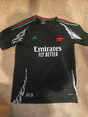 Arsenal x Adidas svart fotbollströja - Säljer en svart Arsenal fotbollströja från Adidas med gröna och röda detaljer på axlarna, vit grafiskt mönster på sidorna och ärmarna samt röd Arsenal-logga på bröstet. Tröjan har Emirates Fly Better-tryck och är tillverkad i lätt polyester.