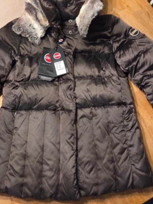 Mörkbrun Colmar dunjacka med fuskpäls - Snygg mörkbrun dunjacka från Colmar med fluffig fuskpäls i huvan och coola detaljer som knappar och dragkedja. Jackan har ett quiltat mönster och långärmade ärmar, perfekt för kalla dagar. Märkeslogga på ärmen och riktigt skön passform.