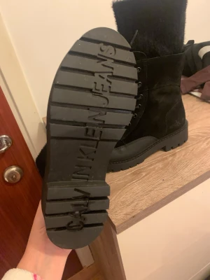 Svarta boots från Calvin Klein Jeans - Säljer ett par svarta boots från Calvin Klein Jeans med grov sula och snörning framtill. Skorna är i mocka med detaljer i skinn och har fluffig kant upptill. Perfekta för höst och vinter, med cool och robust look.