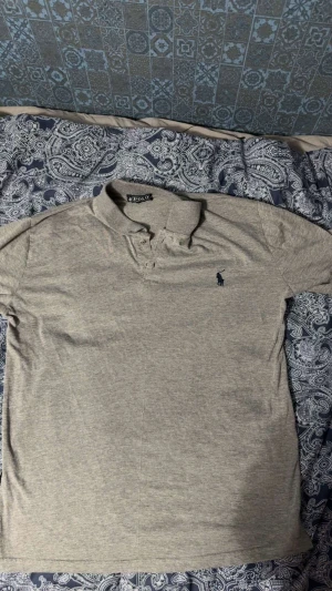 Grå pikétröja från Polo Ralph Lauren - Säljer en stilren grå pikétröja från Polo Ralph Lauren med klassisk krage och knappslå. Tröjan har korta ärmar och en liten mörkblå logga broderad på bröstet. Perfekt för dig som gillar enkel och snygg design.