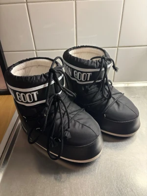 Svarta Moon Boots  - Moon Boots i svart med vit detalj och tydlig logga framtill. Skorna har snörning och en rejäl, räfflad sula för extra grepp. Fodrade på insidan för att hålla värmen och tillverkade i syntetmaterial. Perfekta för kalla vinterdagar och snöäventyr.