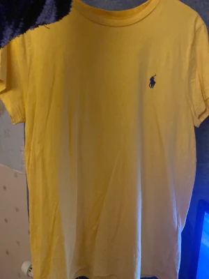 Gul t-shirt från Ralph Lauren - Säljer en gul t-shirt från Ralph Lauren med klassisk blå broderad logga på bröstet. T-shirten har rund hals och korta ärmar, tillverkad i mjuk bomull som är skön mot huden. Perfekt för dig som gillar stilrena och enkla plagg med en touch av ikonisk design.