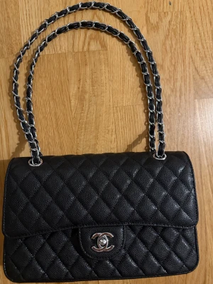 Caviar väska  - En sofistikerad axelväska i strukturerat caviar-läder som kombinerar klassisk elegans med modern funktion. Väskan har ett stilrent quiltat mönster som ger ett exklusivt uttryck och samtidigt gör lädret extra slitstarkt och tåligt för vardaglig användning.  Den silverfärgade kedjeremmen, flätad med läder, ger en lyxig känsla och kan bäras både på axeln eller i handen. Väskans lock stängs med ett dekorativt vridlås i metall som ger ett diskret men ikoniskt intryck.