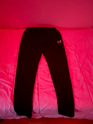 Svarta mjukisbyxor från Under Armour - Svarta mjukisbyxor från Under Armour med logga tryckt på vänster lår. Byxorna har en enkel, rak passform och är gjorda i mjukt syntetmaterial som är perfekt för träning eller chill. Elastisk midja för skön passform och stilren look.