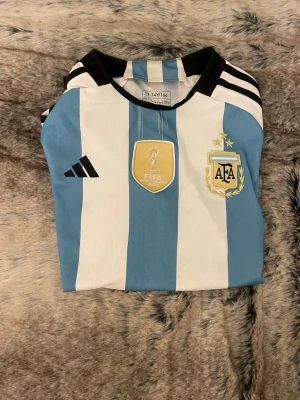 Argentina fotbollströja Adidas 2022 XS - Säljer en officiell Argentina fotbollströja från Adidas, VM 2022. Tröjan har klassiska blå och vita ränder, svarta detaljer och broderade märken på bröstet. Tillverkad i lätt polyester med AFA-emblem och FIFA-märke. Perfekt för fotbollsfans!