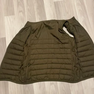 Olivgrön pufferjacka med dragkedja - Säljer en olivgrön pufferjacka med horisontella quiltade paneler och dragkedja. Jackan har en clean look och är perfekt för kyliga dagar. Materialet är syntet och jackan har en modern, loose passform som ger en avslappnad vibe. Knappast använd👍